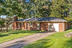 A single-story brick house with a dark shingled roof and an attached garage. The house is surrounded by green grass and trees, with a driveway leading up to the garage. The front yard features a for sale sign.