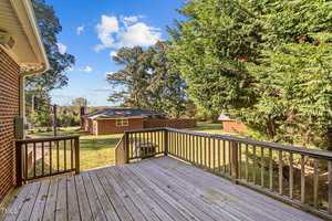 Wooden deck with railing overlooking a grassy backyard. A brick building and a small shed are visible in the distance. The area is surrounded by tall trees, and the sky is partly cloudy.