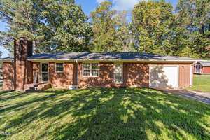 A single-story brick house with a front porch, a chimney, and an attached white garage door. The lawn is grassy with some trees in the background. The sky is clear and sunny.