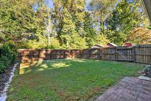 A fenced backyard featuring a grassy lawn bordered by a wooden fence. There are wooded areas with tall trees in the background. In the foreground, a small section of a red brick patio is visible on the right side.