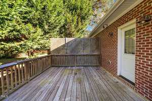 A wooden deck with a railing is attached to a red brick house. A white door leads into the house. A tall lattice privacy screen separates the deck from the surrounding greenery.