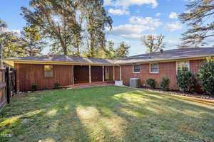 A backyard features a well-maintained lawn bordered by a fence. The single-story brick and wood house has multiple windows, a small porch, and an air conditioning unit on the side. Tall trees are in the background under a partly cloudy sky.