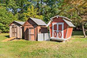 Three wooden sheds in a grassy area are shown. The middle shed is brown with a sloped roof. The shed on the right is red with white trim and double doors. Trees and bushes are visible in the background.