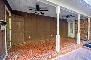 A covered porch features a mosaic tile floor, wooden panel walls, and a white ceiling with two ceiling fans. One wooden door is on the left, and a glass-paneled door is on the right. There are two white pillars supporting the ceiling.