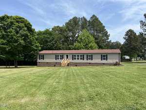 A single-story, rectangular manufactured home at 1686 Briggs Road in Henderson boasts a brick foundation, light siding, and a brown roof. Situated on a spacious, grassy lawn, it has a set of stairs leading to the entrance. The backdrop features tall trees and a clear sky.