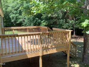A wooden deck with railings, attached to a house, surrounded by trees and grass. Stairs on the right lead down to the ground. Sunlight filters through the leaves, casting shadows on the deck.