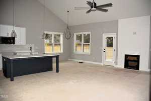 A spacious, unfurnished living room with an open kitchen. Features include gray walls, a high ceiling with a black ceiling fan, a white fireplace, a kitchen island with a sink, pendant lights, and large windows offering natural light.