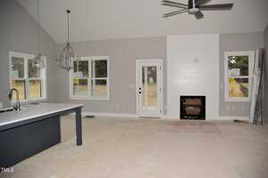 A modern, unfurnished living room with light gray walls and an unfinished floor. It features a fireplace, dark blue kitchen island, large windows, a ceiling fan, and a chandelier. The room has a high, vaulted ceiling and a glass door leading outside.