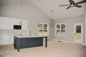 A spacious, unfurnished kitchen and dining area with light gray walls and a high ceiling. It features a dark blue island with a sink, white cabinets, a pendant light, a chandelier, and a ceiling fan. Large windows offer views of a fenced yard.