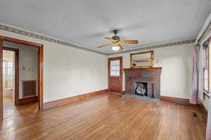 An empty living room with a wooden floor and a brown brick fireplace. A ceiling fan with a light is mounted on the ceiling. There is a mirror above the fireplace, and a wooden door with a window. Windows with curtains are on the right wall.