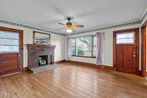 A living room with wooden floors and a brick fireplace. A ceiling fan is centered above. The room has two wooden doors and a large window with sheer curtains, allowing natural light inside. A mirror hangs above the fireplace.