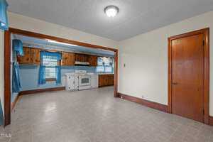 A spacious kitchen and dining area with wooden cabinets and blue curtains. The room features a white stove and range hood, tiled flooring, and wood-paneled accents. A wooden door and wide doorway connect the two areas. A single overhead light fixture is present.