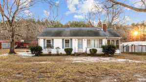 A small, single-story white house sits peacefully along 2677 Nc 39 Hwy S in Henderson. It features a dark roof and a welcoming front porch. The yard is grassy with patches of dirt, complemented by a few bushes in front and a large tree to the right, as the sun sets behind distant trees.