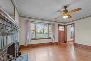 A spacious living room with a wooden floor, brick fireplace, and ceiling fan. The room has large windows with curtains, allowing natural light. A wooden door with glass panels is visible. The walls are light-colored with decorative trim at the top.