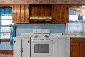 A kitchen with a vintage design featuring wooden cabinets and drawers. An electric stove with four burners is centered between the cabinets. The backsplash is a light blue color, and the windows have blue and white patterned curtains.