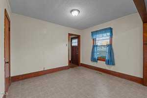 An empty room with beige walls and gray tile flooring. A window with blue curtains is on the right, and a wooden door to the outside is at the back. A light fixture is on the ceiling, and wooden trim borders the floor and doorway.