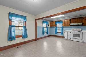 An empty room with a tiled floor features two windows with blue curtains. The room is partially divided by a wooden frame, leading to a small kitchen area with wooden cabinets. A white oven and range are positioned against a wall under a fluorescent light.