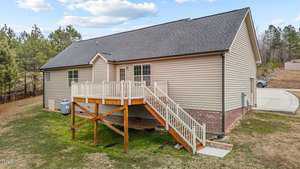 A single-story house with beige siding and a dark gray roof. There is a wooden deck with a staircase leading to the grass below. The house is surrounded by trees, and a concrete driveway is visible on the right side.