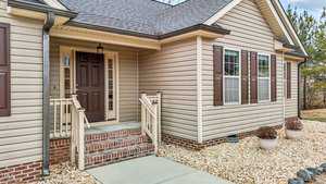 A beige house exterior with a front porch featuring a brown door and steps with railings. The porch is brick-lined at the base. There are two windows with brown shutters, and the yard is landscaped with white stones and potted plants.