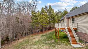 A wooden deck with stairs leads down to a grassy backyard of a house. The house is surrounded by a mix of trees, some without leaves and some evergreen. The sky is partly cloudy.