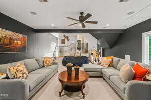 A modern living room with gray walls and a sectional sofa adorned with colorful throw pillows. A round wooden coffee table sits in the center. A ceiling fan hangs above, and a staircase with a wooden railing leads to an upper level.