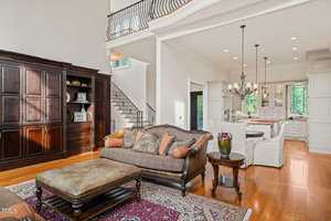 A spacious living room with a brown leather sofa and matching ottoman on a patterned rug. An ornate chandelier hangs above a dining area in the background. A dark wooden armoire is against the wall, and a staircase leads to an upper floor.