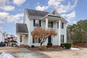 Two-story light blue house with white trim and a front porch supported by columns. A leafless tree is in the front yard. The driveway and yard have patches of snow. The sky is partly cloudy, and other houses are visible in the background.
