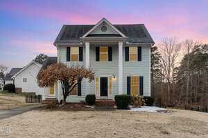 A two-story colonial-style house at 2047 Ferbow Street in Creedmoor is pictured at sunset. It features light blue siding with white trim, a black shingled roof, symmetrically placed windows with shutters, and a front porch with columns. The lawn is lightly browned, with a tree in front.
