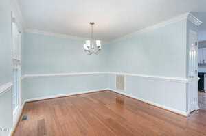 An empty dining room with light blue walls and white trim features a wooden floor. A modern chandelier hangs from the ceiling. Two windows with blinds are present, and a white door leads to another room.
