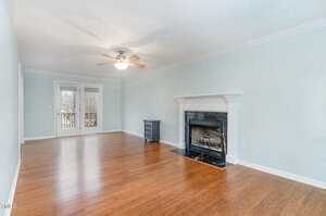 A spacious, empty living room features light blue walls, a ceiling fan, and a fireplace with a black hearth. The floor is hardwood, and glass doors lead to the outside. A small side table stands near the wall.