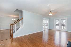 A spacious, empty living room with light blue walls and hardwood flooring. A ceiling fan is mounted above. Wooden stairs with white balusters lead to the upper floor. French doors open to a deck, and a chandelier hangs in the adjacent dining area.