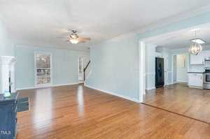 A spacious, empty room with light blue walls and wood flooring. The room features a ceiling fan and large windows. There is an adjacent kitchen area with white cabinets and a black refrigerator visible in the background.