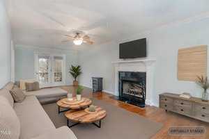 A virtually staged living room with a sectional sofa, two round coffee tables, and a fireplace with a TV mounted above. A ceiling fan is on the white ceiling. The walls are light-colored, and there's a large window and door at the back.