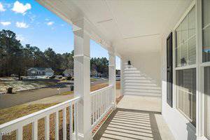 A white porch with a railing and two columns overlooks a quiet suburban street. The area is surrounded by trees under a clear blue sky. A light fixture is attached to the wall near the entrance. The porch floor is concrete.