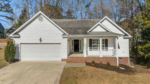 A single-story white house on 2773 Clifton Avenue in Creedmoor, with a gray roof, features a two-car garage and a small front porch with columns. The yard is bordered by a few small shrubs and trees, and there is a paved driveway leading to the garage.