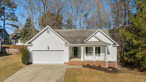 Single-story house with light gray siding and a two-car garage. It has a small front porch with white railings and columns. The driveway leads to the garage, and there are trees in the background and a small lawn in front.