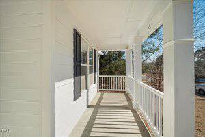 A wooden porch with a white railing and posts is attached to a house with white siding. The porch is covered by a roof, and there is a large window with black shutters on one side. Sunlight casts shadows of the railing on the floor.