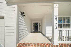 A white house front porch with a black front door featuring a wreath. There's a porch light on the left and the number 2773 displayed on the right column. A window with shutters is partially visible on the right side.