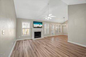 Spacious living room with light wood flooring, high ceiling, and light gray walls. Features a ceiling fan, a fireplace with a TV above, and large windows with plantation shutters. French doors lead to an outdoor area. A chandelier is in the corner.
