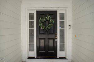 A black front door with a green wreath is centered between two narrow vertical windows. The exterior walls are light-colored with horizontal siding. A doorbell is on the right side of the door.