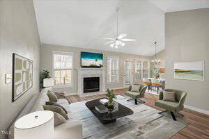 A modern living room featuring a beige sofa, two green armchairs, and a black coffee table on a patterned rug. A fireplace with a TV above it centers the room. Large windows and a dining area with a chandelier are visible in the background.