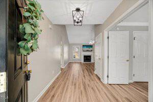 A hallway with light wood flooring and beige walls leads to a living room with a fireplace and ceiling fan. A black lantern-style light fixture hangs from the ceiling, and a door with a wreath is visible on the left.