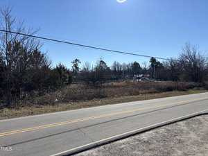 A two-lane road with yellow dividing lines runs alongside a grassy, overgrown area near Old Norlina Road in Henderson. Trees and sparse vegetation are visible in the background under a clear blue sky, while power lines stretch across the scene.