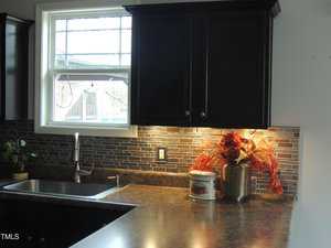 A kitchen counter with a stainless steel sink and a window above. Dark cabinets with a mosaic tile backsplash are visible. The counter holds a potted plant, a tin container, and a vase of orange and red foliage.