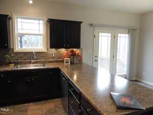 A kitchen with dark cabinets and a marble countertop. There's a sink under a window, a backsplash of small tiles, and a laptop on the counter. A small plant is near the sink. French doors in the background lead outside.