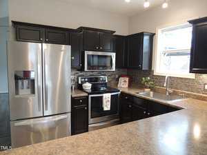 Modern kitchen with dark wood cabinets and gray countertops. Stainless steel appliances include a refrigerator, oven with stove, and microwave. Tiled backsplash in a mosaic pattern. A window above the sink brings in natural light.