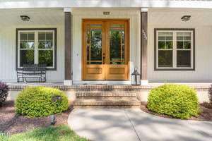 Front porch of a house featuring double wooden doors with glass panels, flanked by two windows. Steps lead up to the porch, and a lantern sits beside the door. Green bushes line the path, and a small bench is on the left side. House number 2323 is visible.