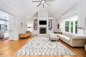 Spacious living room with a high ceiling, featuring a patterned rug, white tufted sofa, beige armchair, and an ottoman. A fireplace with a TV above is centered between built-in shelves. Large window on the right and a view into the kitchen on the left.