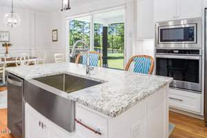 A modern kitchen with a large granite island featuring a stainless steel sink. Behind the island, there are blue and white chairs. A built-in oven and microwave are on the right, and large windows offer a view of trees outside.