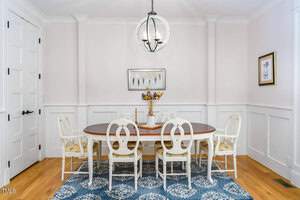 A dining room with a wooden oval table and eight white chairs on a blue patterned rug. A round light fixture hangs above. The walls are white with wainscoting, and there is a framed picture and a vase with dried flowers on the table.